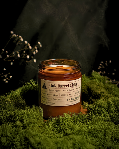 A Farmhouse Charm Candles brand 'Oak Barrel Cider' scented candle in a clear glass jar, resting on a bed of green moss with small white flowers in the background against a dark, moody backdrop.