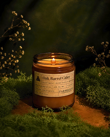A lit amber glass jar candle labeled 'Oak Barrel Cider' by Farmhouse Charm Candles, styled on mossy ground with small white flowers against a dark green background.