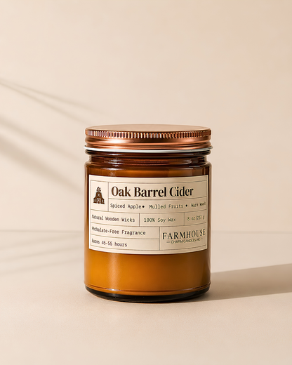 A Farmhouse Charm Candles brand Oak Barrel Cider scented candle in an amber glass jar with a copper-toned metal lid, set against a warm beige background with soft leaf shadows.