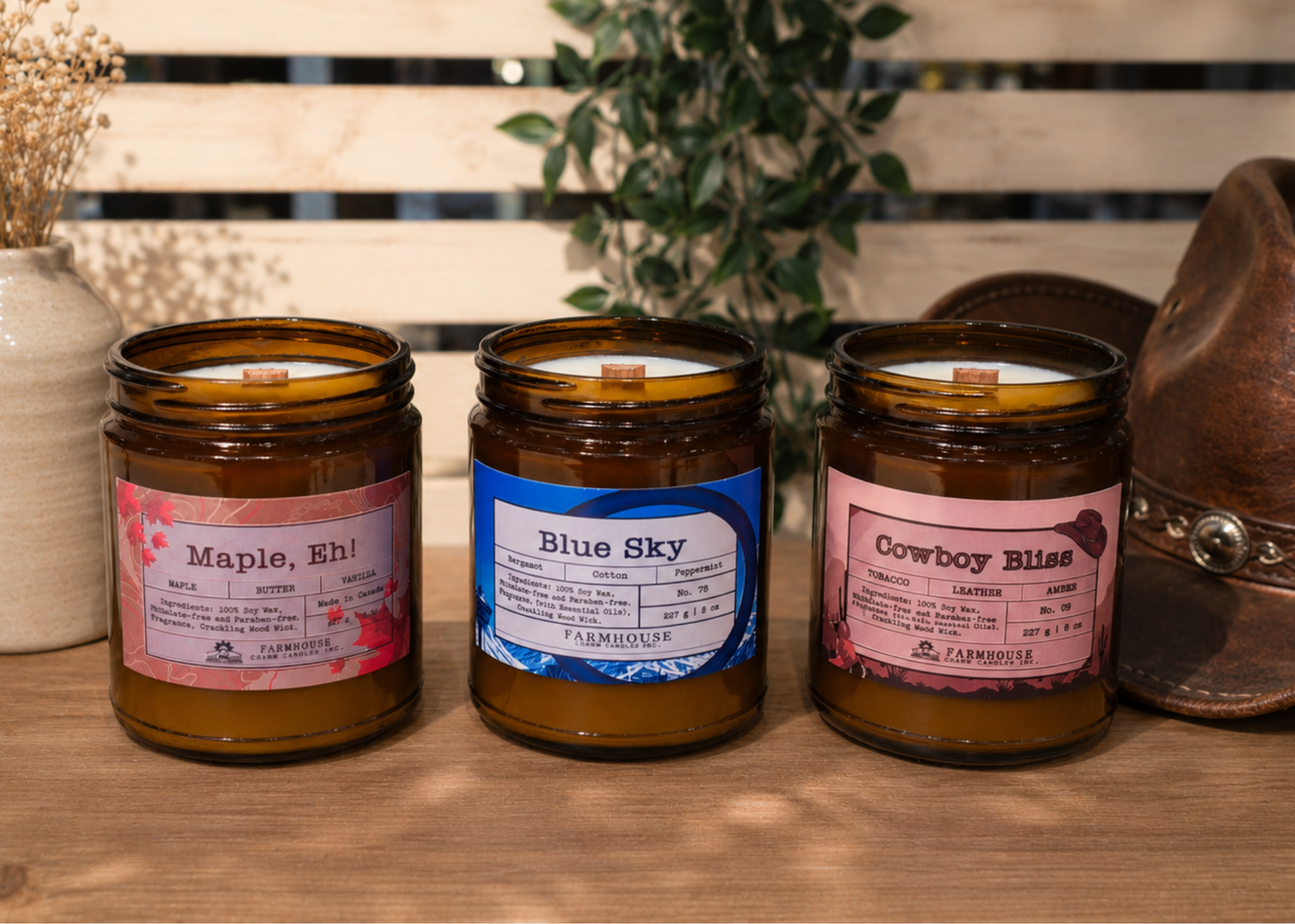 Three jars of candles by Farmhouse Charm Candles with labeled scents (Maple, Eh, Blue Sky and Cowboy Bliss  on a wooden and western themebackground.
