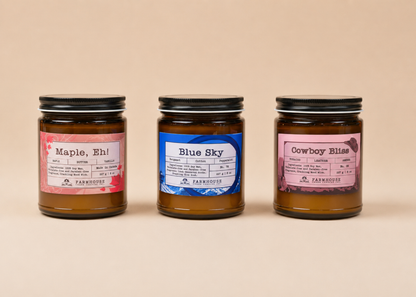 Three jars of candles by Farmhouse Charm Candles with labeled scents (Maple, Eh, Blue Sky and Cowboy Bliss  on a beige background