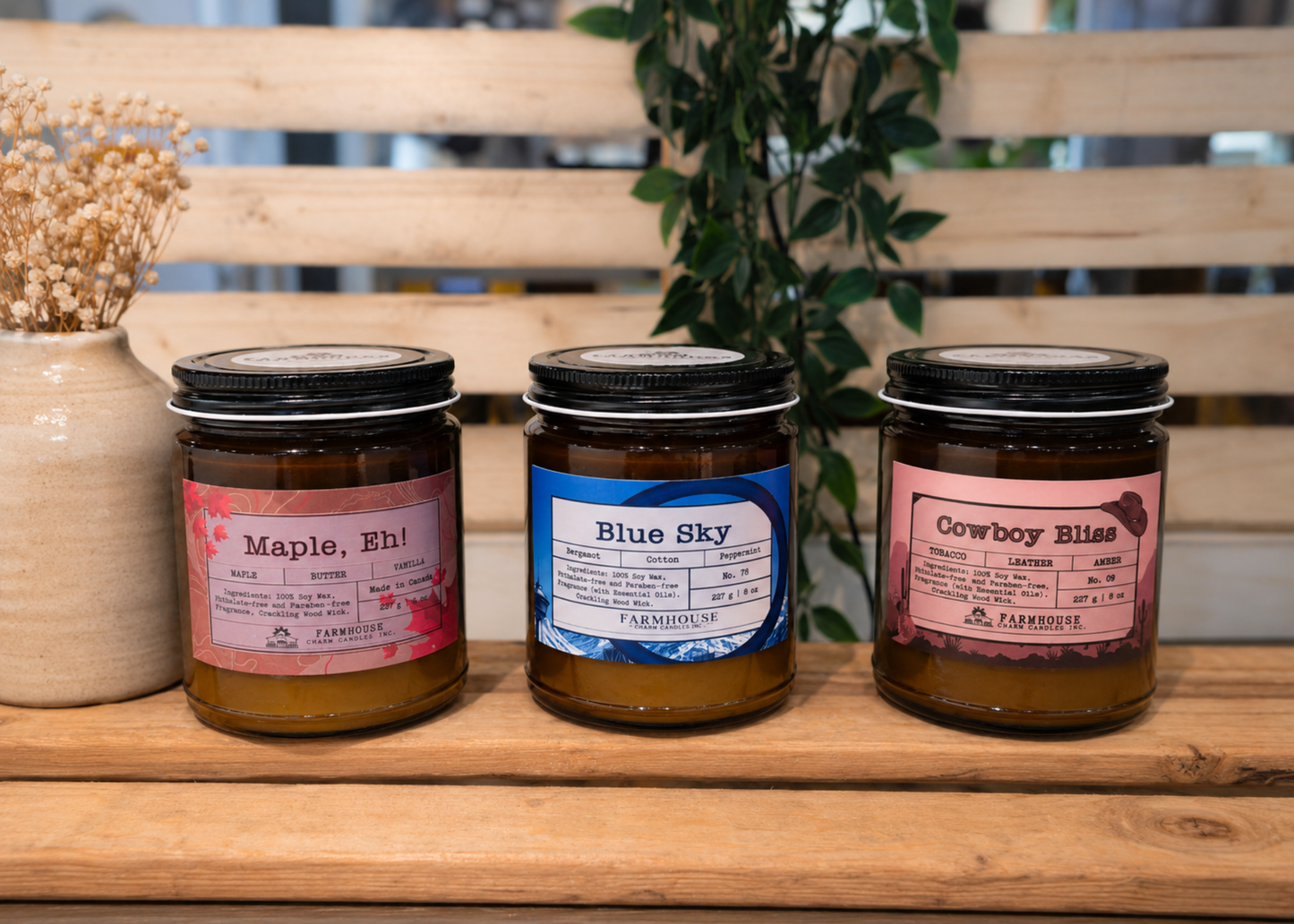 Three jars of candles by Farmhouse Charm Candles with labeled scents (Maple, Eh, Blue Sky and Cowboy Bliss  on a wooden and plant background.
