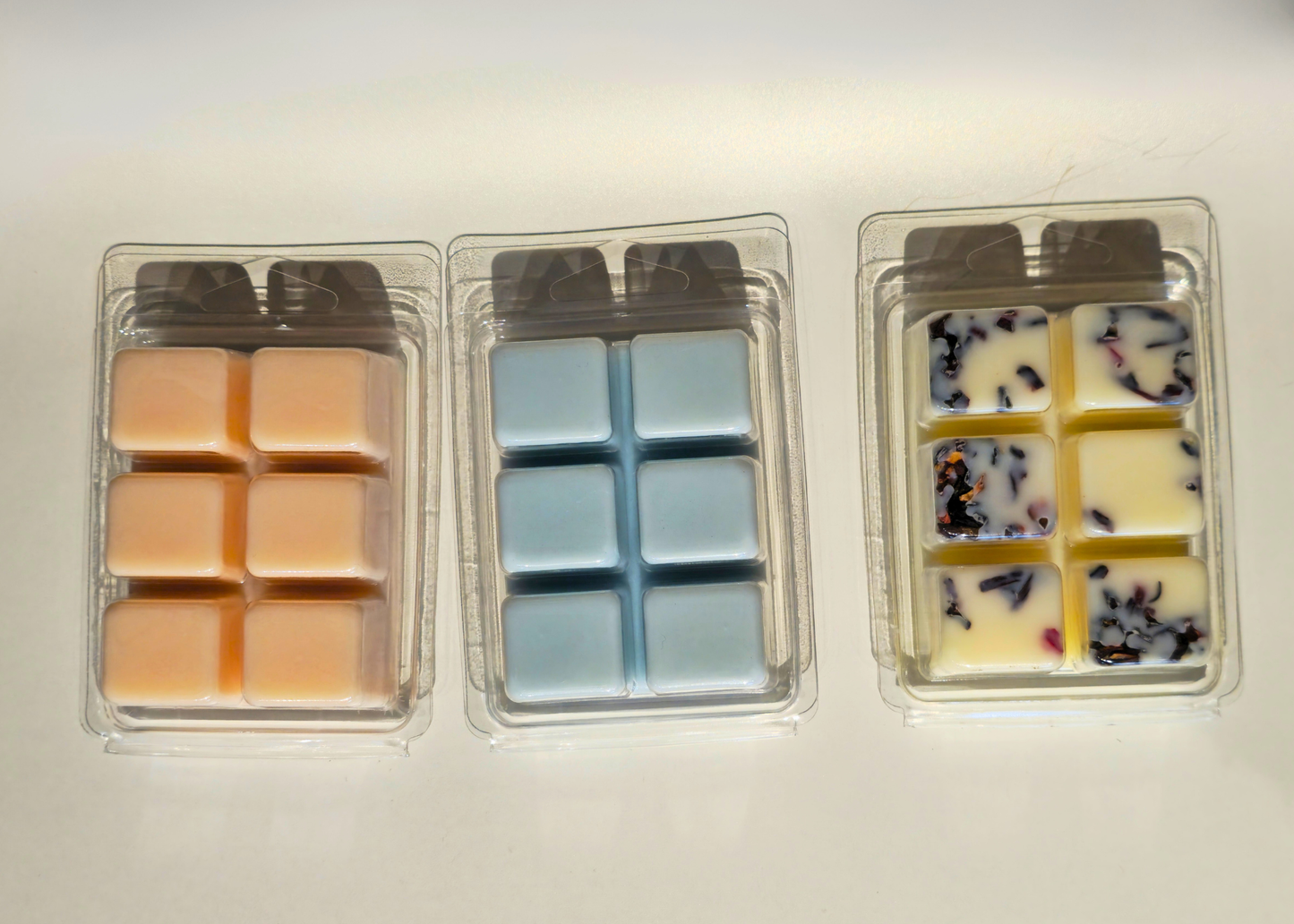 Three clear plastic containers with different colored wax melts on a light background