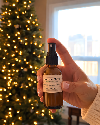 A hand holding a small amber glass spray bottle labeled 'Peppermint Mocha' by Farmhouse Charm Candles, with a blurred Christmas tree and bright window in the background.