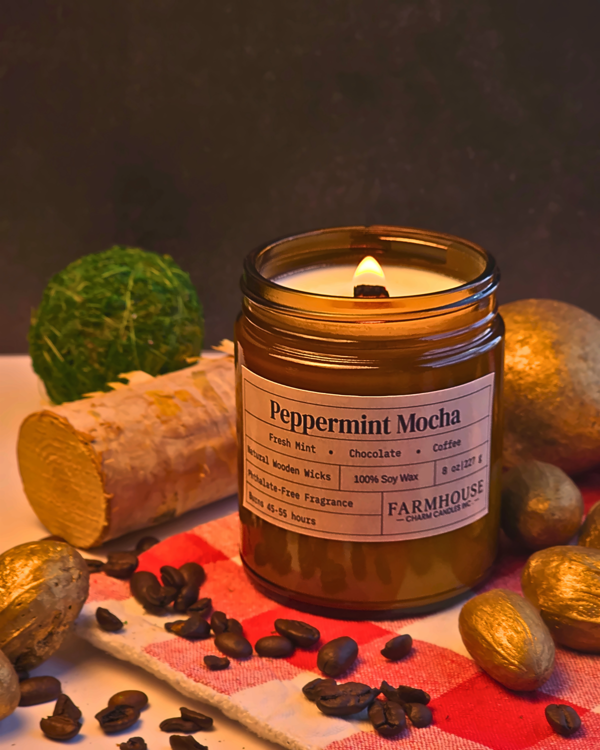 A lit amber glass jar candle labeled 'Peppermint Mocha' by Farmhouse Charm Candles styled with coffee beans, a ginger root, and gold leaf on a red-and-white checkered cloth against a dark background.