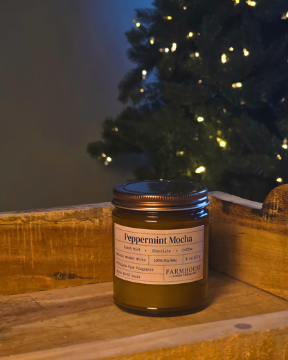A glass jar candle labeled 'Peppermint Mocha' by Farmhouse Charm Candles  placed on a rustic wooden surface with a softly blurred Christmas tree in the background.