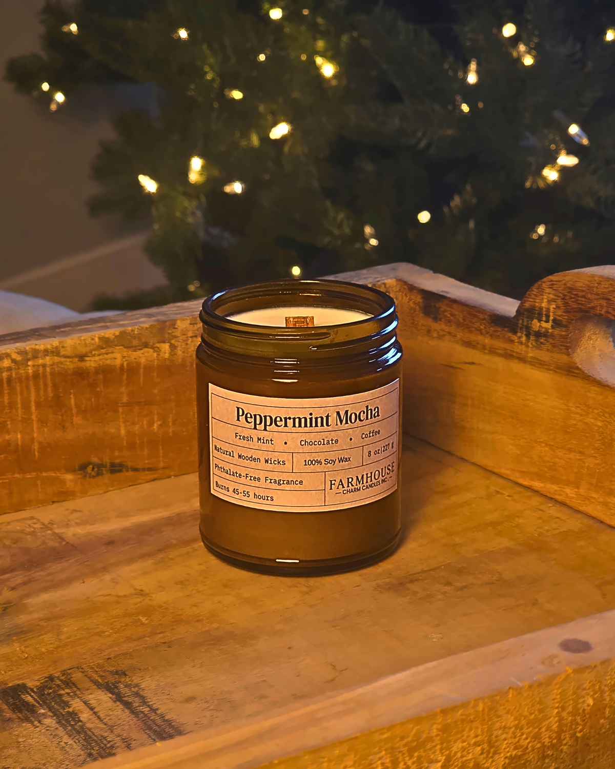 A Farmhouse Charm Candles  in an amber glass jar sits on a rustic wooden surface with a blurred Christmas tree in the background.