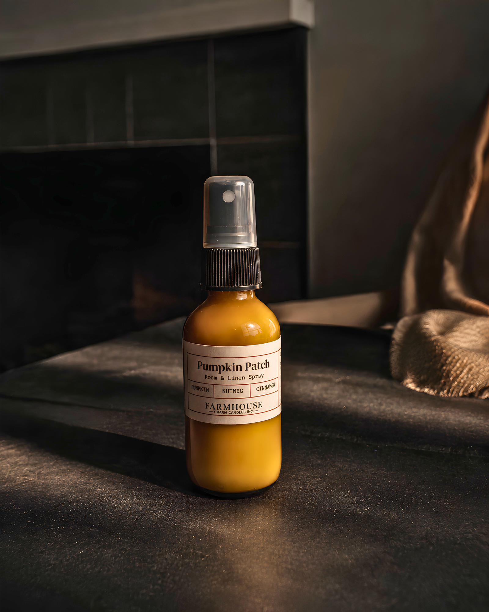 A small amber spray bottle labeled 'Pumpkin Patch' by  Farmhouse Charm Candles, filled with  liquid, set on a dark textured surface with warm, moody lighting.