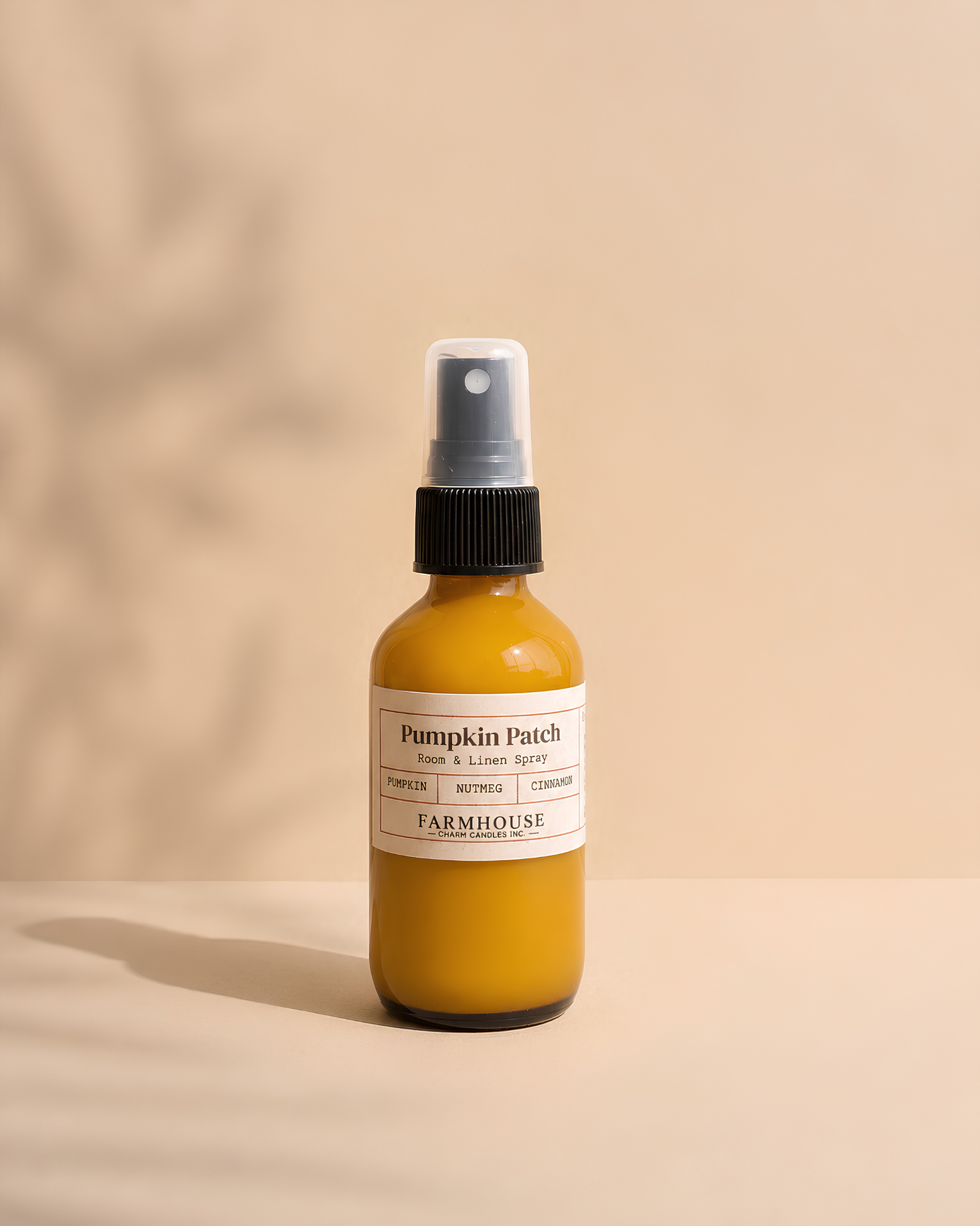 A small amber glass spray bottle labeled 'Pumpkin Patch' by Farmhouse Charm Candles, filled with liquid, standing on a beige surface against a matching beige background with soft leaf shadows.