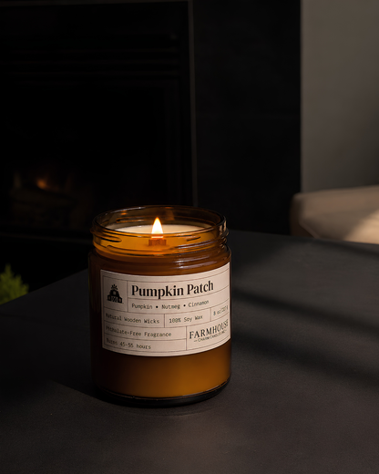 A lit amber glass jar candle labeled 'Pumpkin Patch' sits on a dark tabletop in a dim indoor setting. The label also shows a brand name Farmhouse Charm Candles