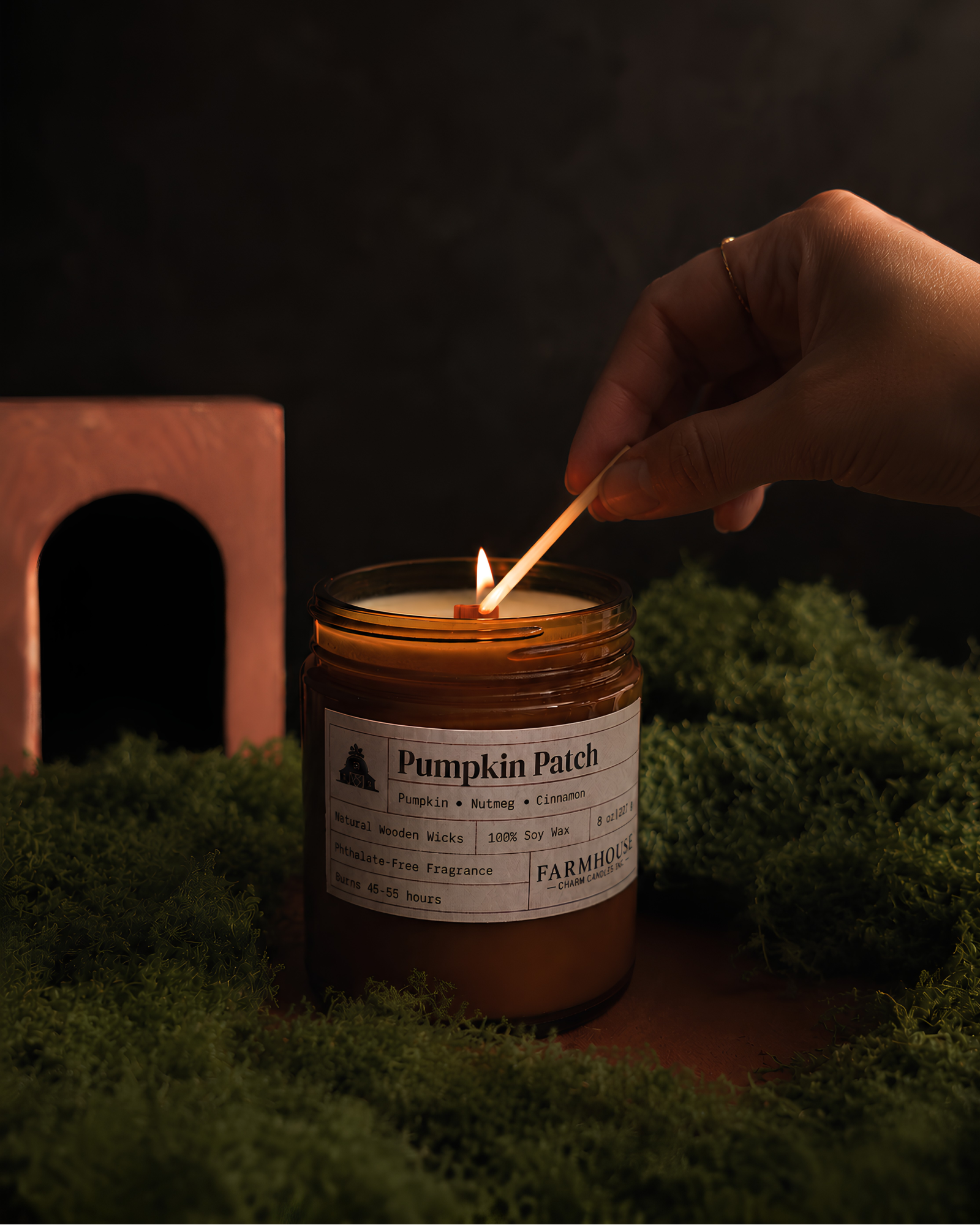 A lit amber glass jar candle labeled 'Pumpkin Patch' by Farmhouse Charm Candles being lit with a wooden match by a hand, set on green moss with a rustic terracotta arch in the background.