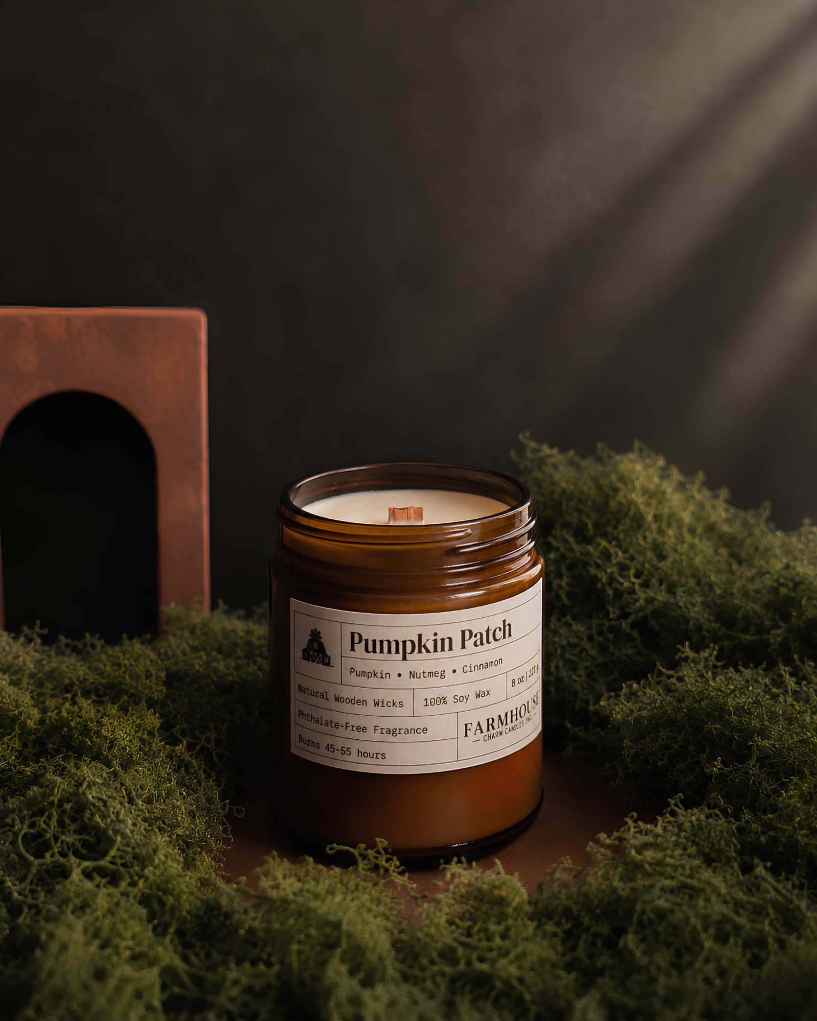 A jar candle labeled 'Pumpkin Patch' by Farmhouse Charm Candles, set in green moss with a dark textured background and a wooden block prop.