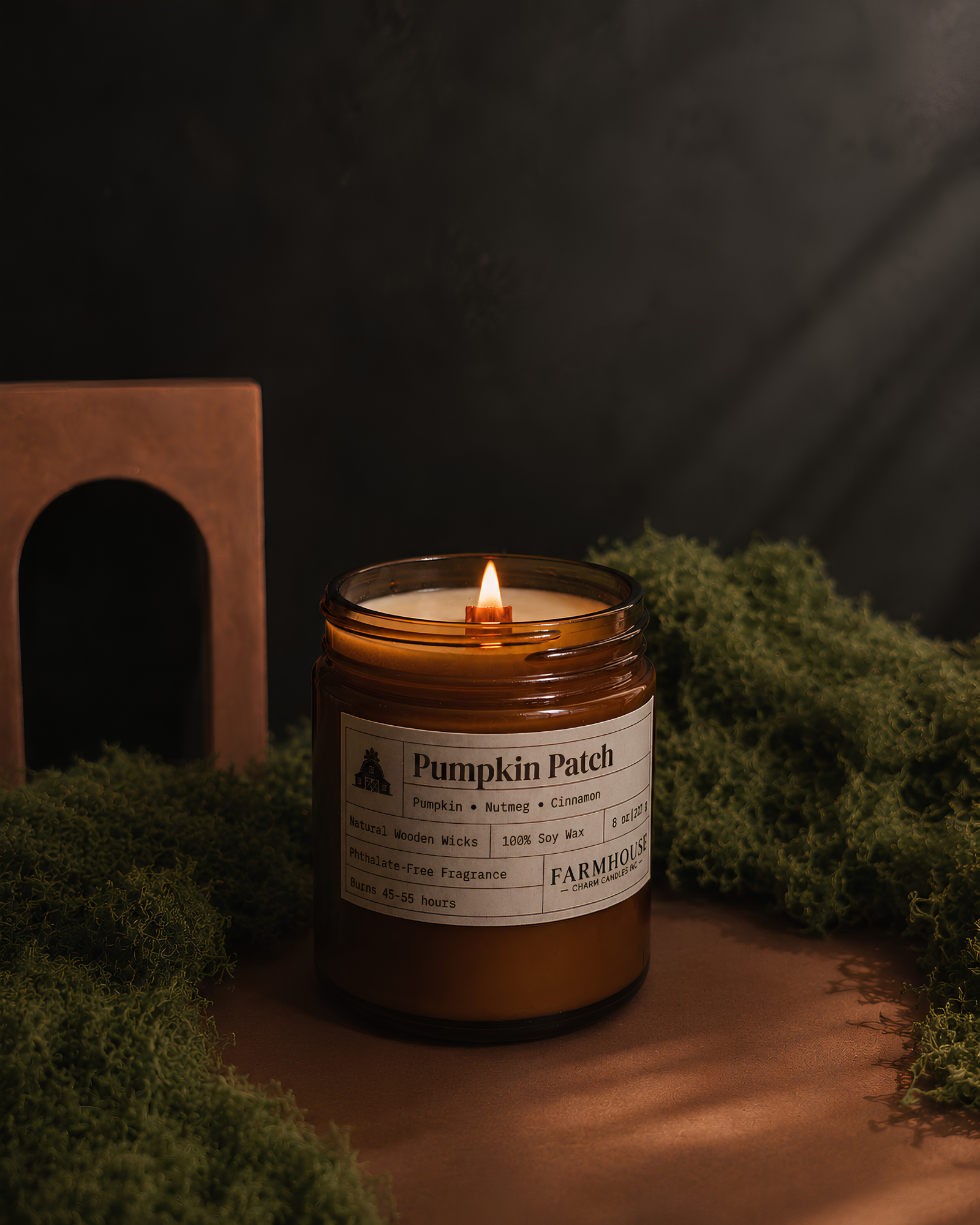 A lit amber glass jar candle labeled 'Pumpkin Patch' by Farmhouse Charm Candles, styled on a wooden surface with green moss and a dark textured background.