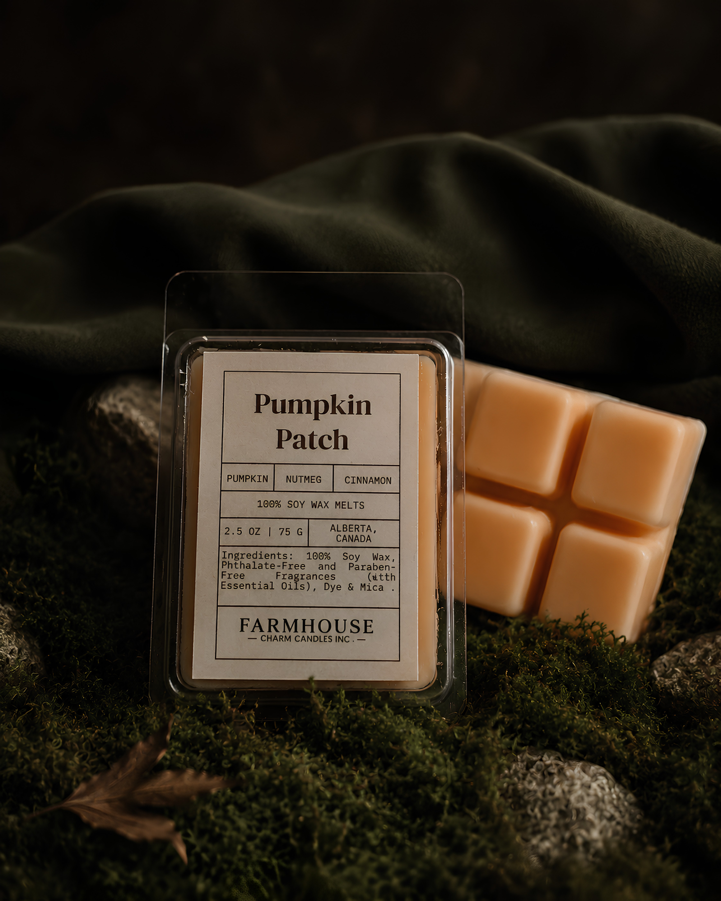 A packaged pumpkin patch scented wax melt from Farmhouse Charm Candles , displayed next to a partially melted wax cube on a bed of dark green moss and fabric.