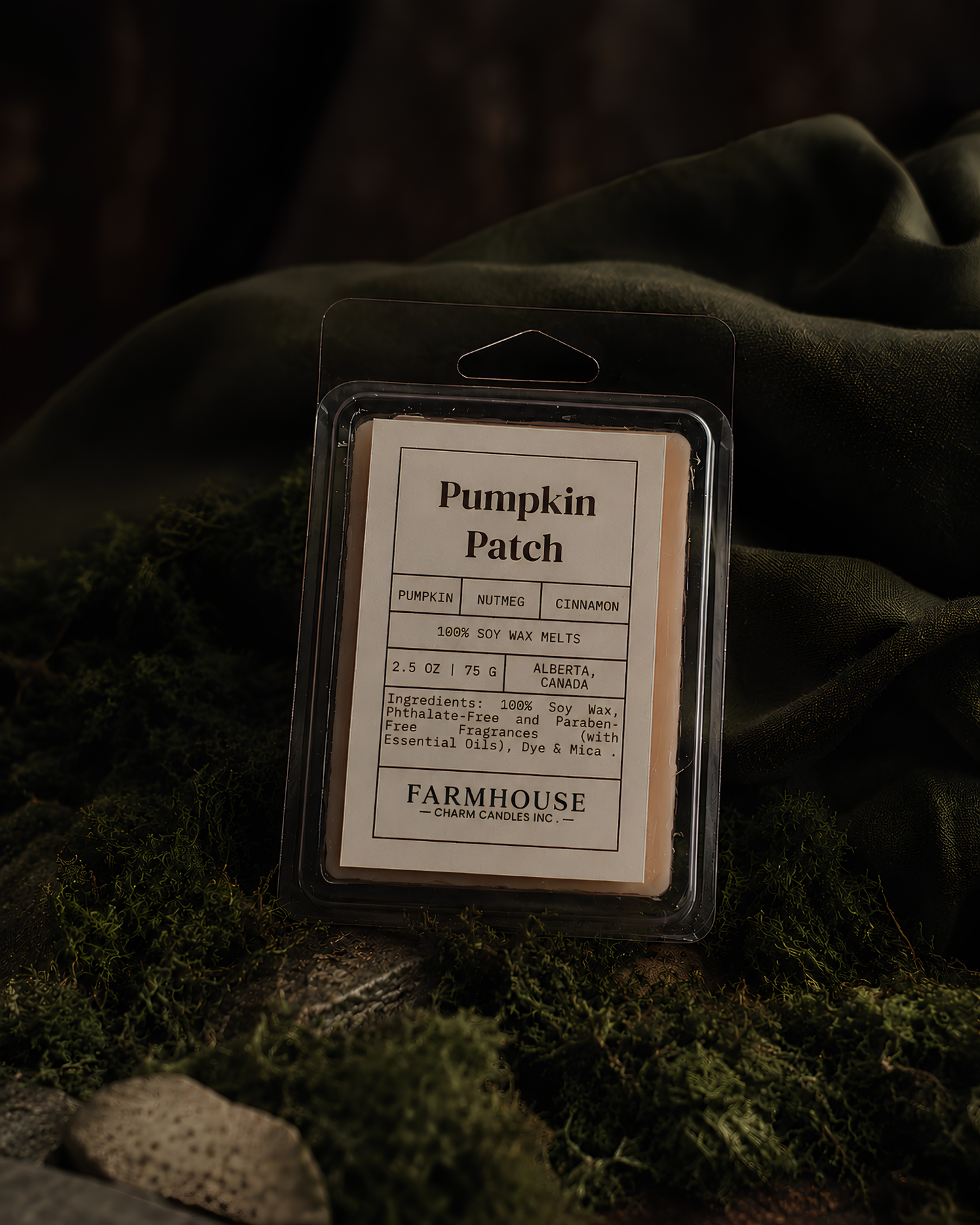 A packaged wax melt labeled 'Pumpkin Patch' by Farmhouse Charm Candles Company, shown in a clear plastic clamshell with a cream paper label, resting on dark green fabric and moss.