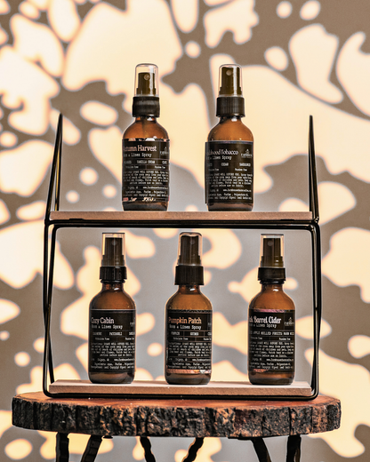 Set of five brown room & linen spray bottles by Farmhouse Charm Candles with labels on a wooden shelf against a leafy background