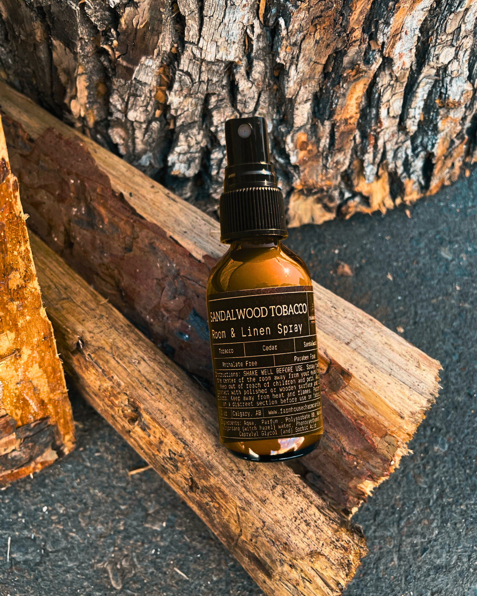 Bottle of Sandalwood Tobacco Room & Linen Spray by Farmhouse Charm Candles on wooden logs with a tree bark background