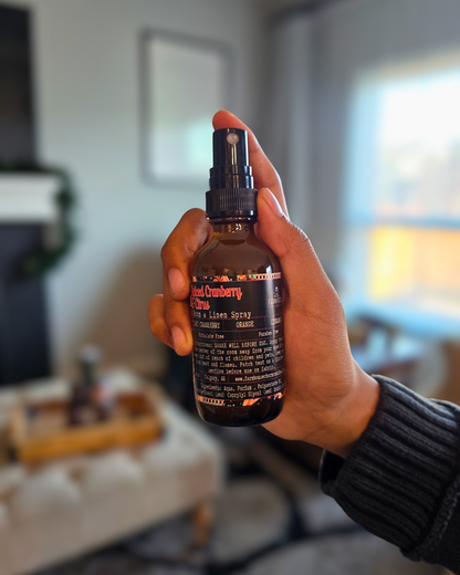 Hand holding an amber glass bottle of Spiced Cranberry & Citrus Room and Linen Spray — evokes a warm, cozy Christmas scent of cranberry, orange, and cinnamon.