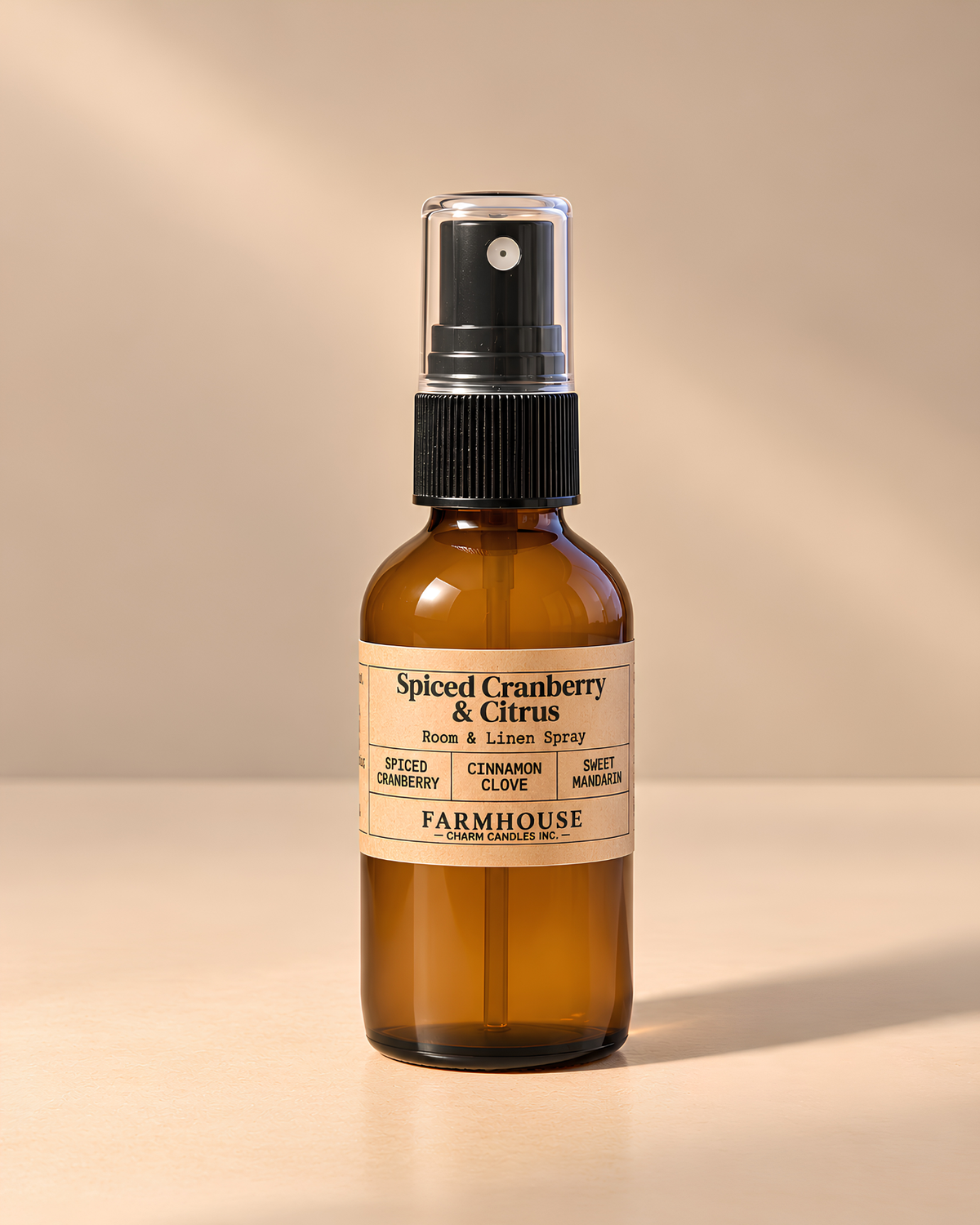 An amber glass room and linen spray bottle with a black spray cap and a cream label reading 'Spiced Cranberry & Citrus' by 'FARMOUSE Charm Candles', photographed on a warm beige background with soft shadows.