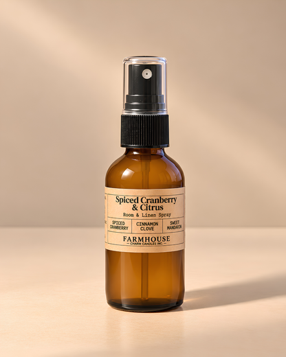 An amber glass room and linen spray bottle with a black spray cap and a cream label reading 'Spiced Cranberry & Citrus' by 'FARMOUSE Charm Candles', photographed on a warm beige background with soft shadows.
