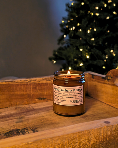 A lit jar candle labeled 'Spiced Cranberry & Citrus' by Farmhouse Charm Candles sits on a rustic wooden surface with a blurred Christmas tree in the background.