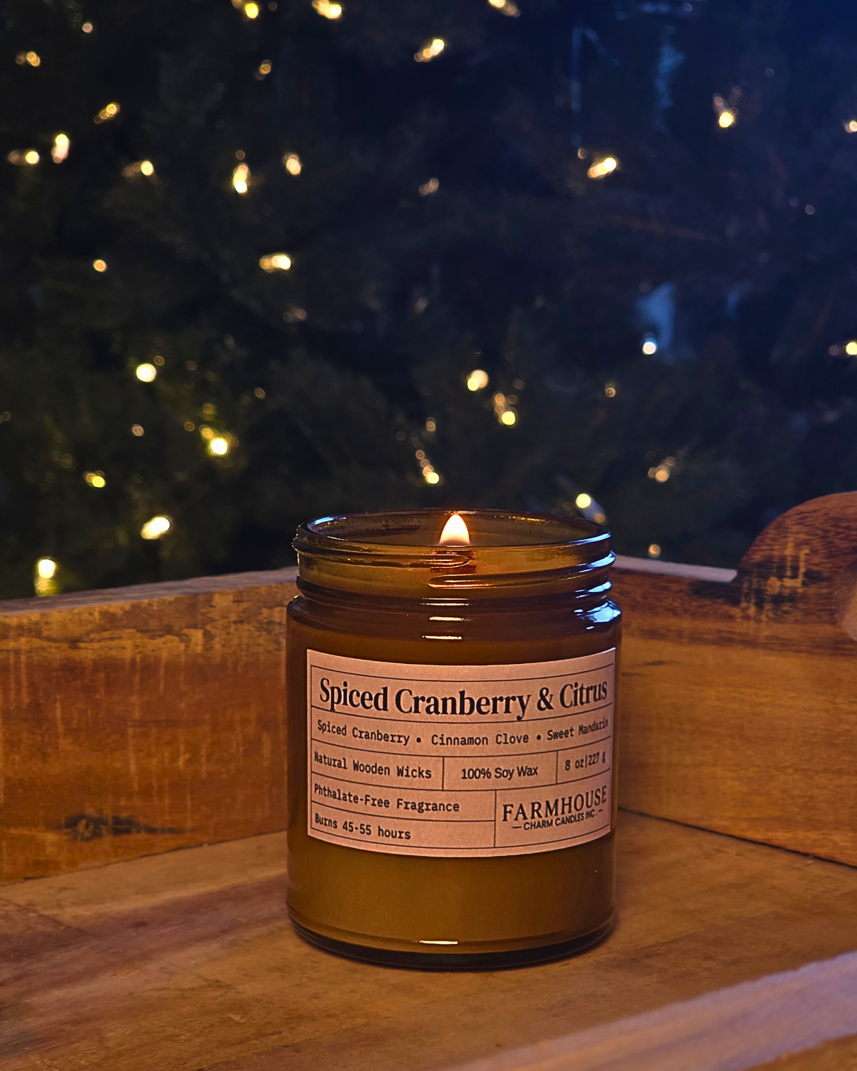 A lit amber glass jar candle labeled 'Spiced Cranberry & Citrus' by Farmhouse Charm Candles placed on a wooden surface with a blurred background of warm string lights and dark foliage.