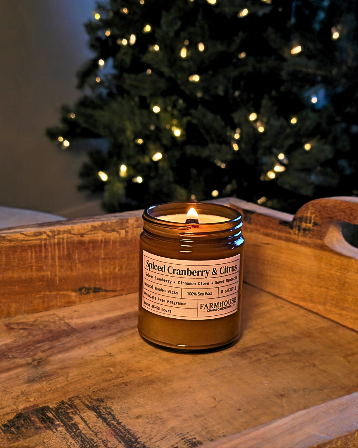 A lit spiced cranberry & citrus scented candle in an amber glass jar sits on a rustic wooden tray, with a blurred Christmas tree in the background. Farmhouse Charm Candles