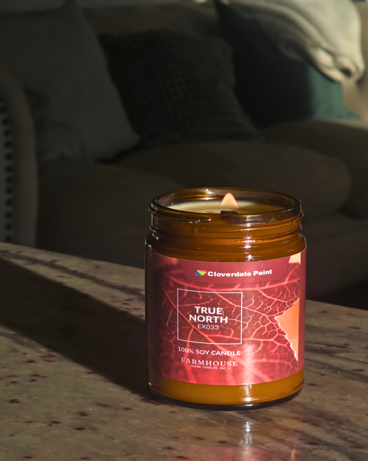 A lit amber glass jar candle sits on a marble countertop in a dimly lit living room. The candle has a pink and red leaf-patterned label with the brand name Cloverdale Paint and the scent name True North by Farmhouse Charm Candles
