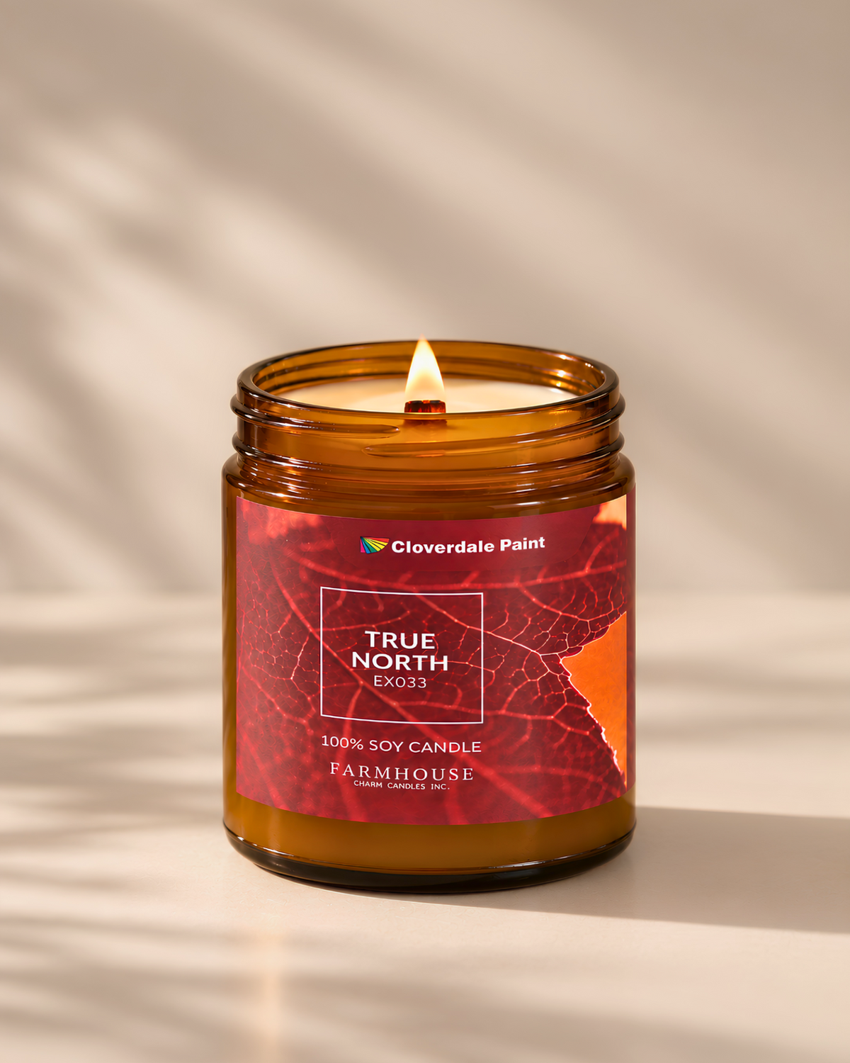 A lit amber glass jar candle with a red and orange leaf-patterned label reading 'TRUE NORTH EX033' and 'Cloverdale Paint,' set on a warm beige surface with soft leaf shadows. Farmhouse Charm Candles