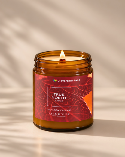 A lit amber glass jar candle with a red and orange leaf-patterned label reading 'TRUE NORTH EX033' and 'Cloverdale Paint,' set on a warm beige surface with soft leaf shadows. Farmhouse Charm Candles