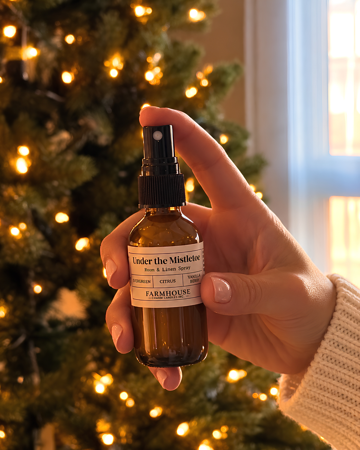 A hand holding a small amber glass spray bottle labeled 'Under the Mistletoe' by Farmhouse Charm Candles, set against a softly blurred Christmas tree with warm lights.