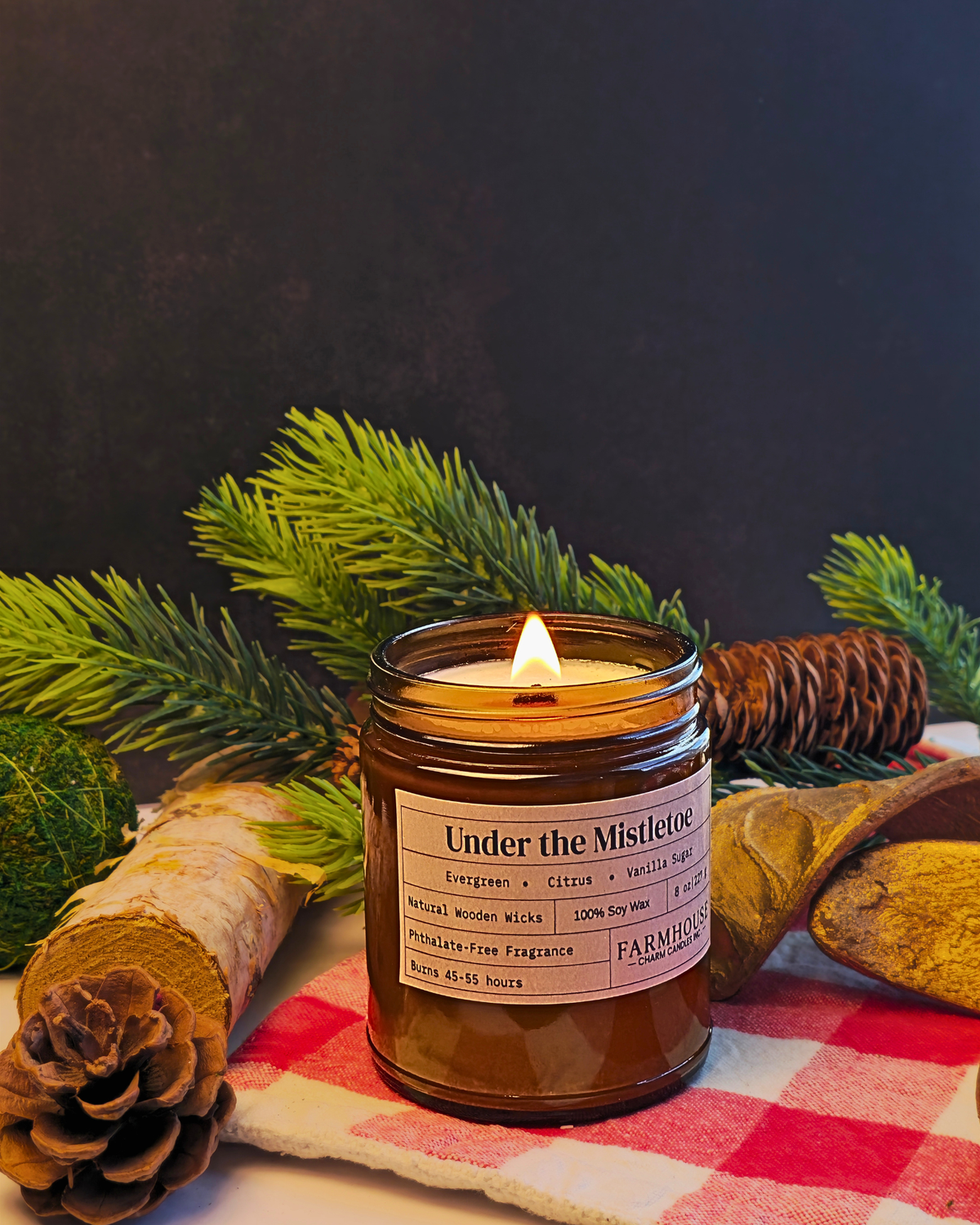 A lit scented candle in a brown glass jar sits on a red-and-white checkered cloth with pine branches, a pinecone, and wood pieces around it against a dark background. The label reads 'Under the Mistletoe' by Farmhouse  Charm Candles.