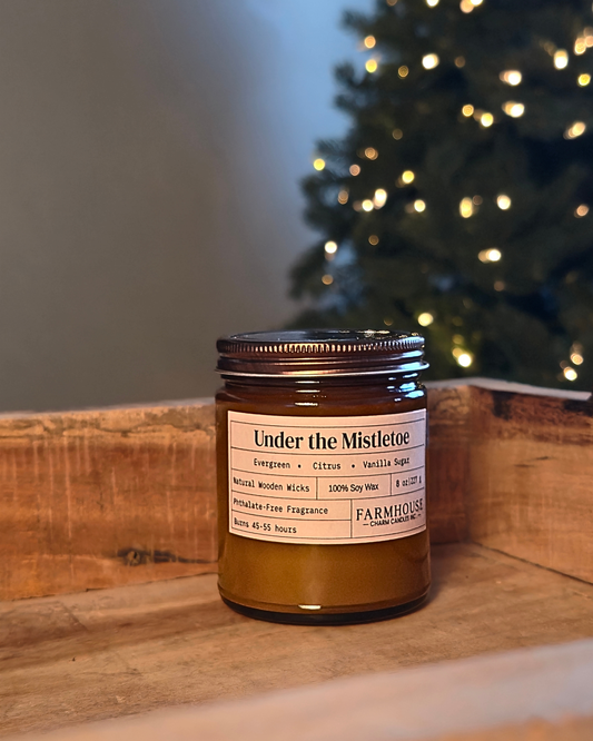 An amber glass jar candle with a metal lid and a cream label reading 'Under the Mistletoe'  by Farmhouse  Charm Candle sits on a rustic wooden ledge with a blurred Christmas tree in the background.