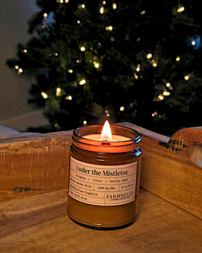 A lit  Farmhouse  Charm Candle 'Under the Mistletoe' soy candle in an amber glass jar, set on a rustic wooden surface with a blurred Christmas tree in the background.