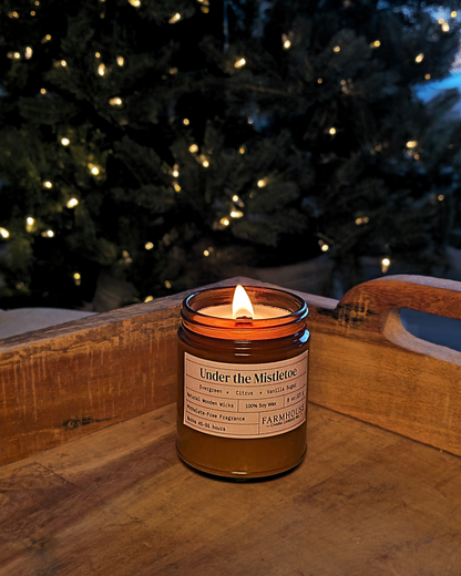 A lit amber glass jar candle labeled 'Under the Mistletoe' by Farmhouse  Charm Candles placed on a wooden tray with a blurred Christmas tree of warm lights in the background.